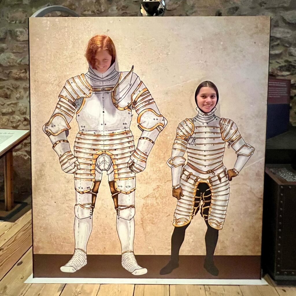 A photo booth backdrop featuring two individuals in detailed plate armor, displayed on a wooden floor inside a stone structure.