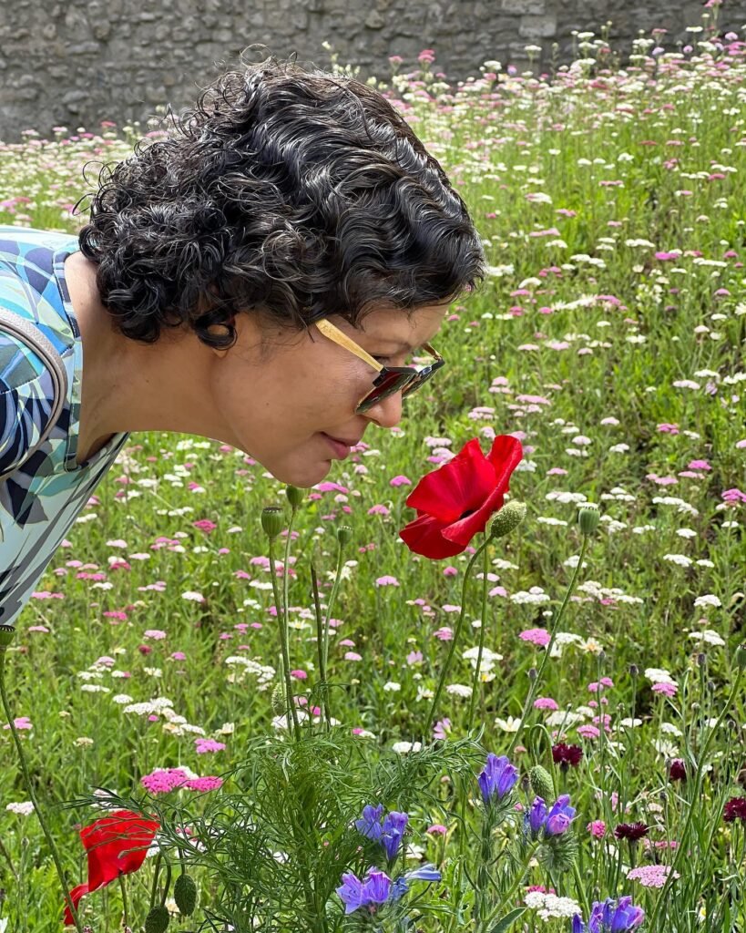 A person leans down to inspect a red flower in a field of colorful wildflowers, with closeups of curly hair and colorful blooms.