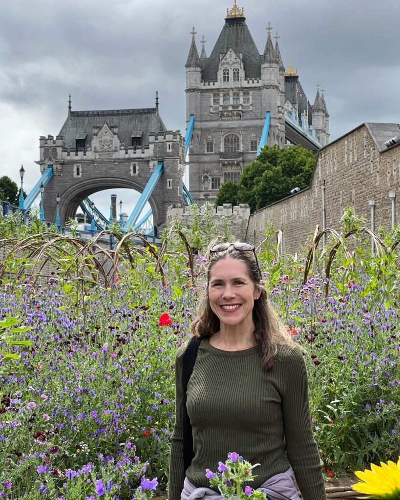 A person stands smiling in a garden with purple flowers while Tower Bridge looms in the background under a cloudy sky.