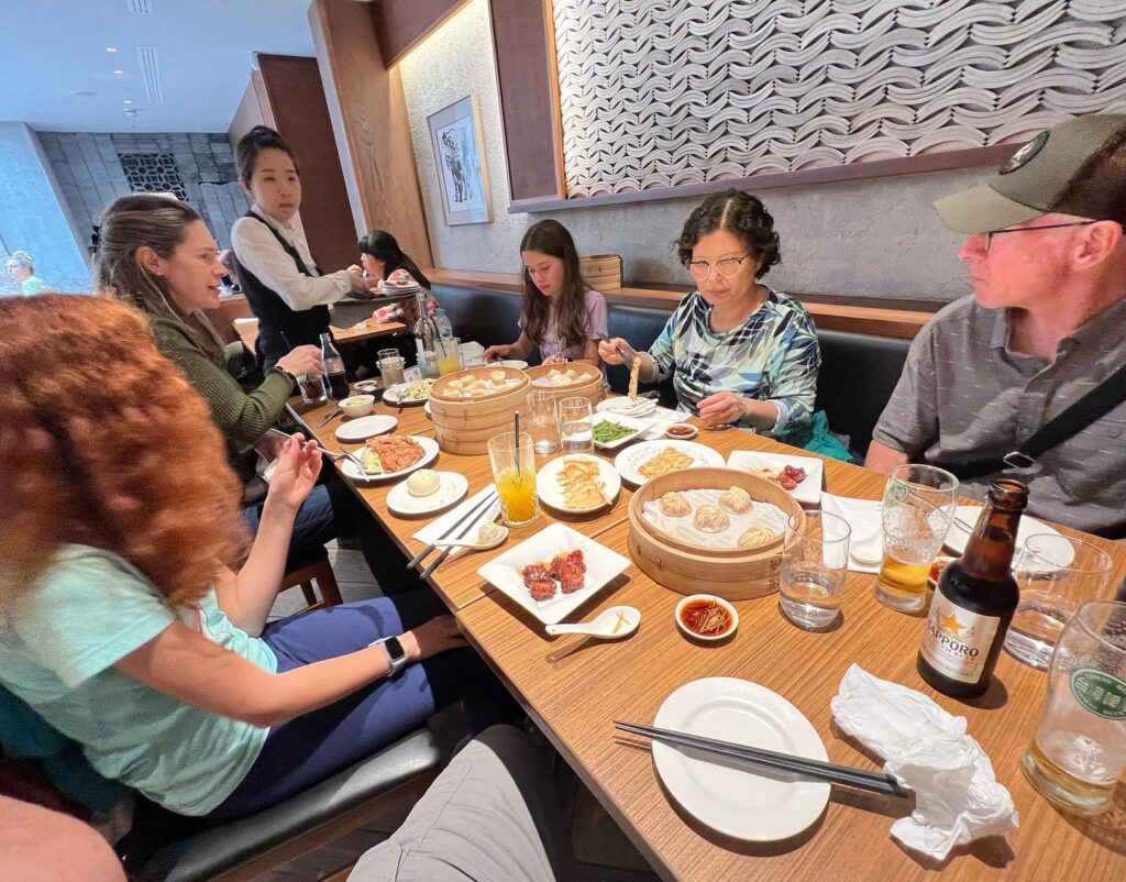 A group of people enjoying dim sum and drinks at a restaurant. The table is set with baskets of dumplings and various dishes.