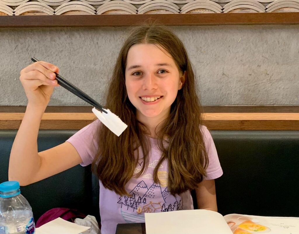 A child is seated at a restaurant booth, smiling while holding chopsticks and a small object. A water bottle sits on the table nearby.