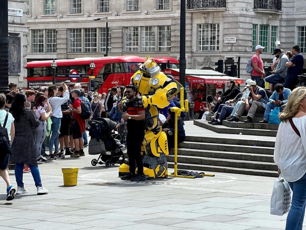 A man in a Big Bird costume poses for photos in a London square. Red double-decker buses and crowd of people in the background.