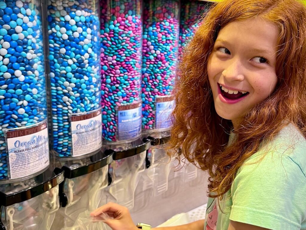 A view of a colorful self-serve candy machine filled with blue and pink sweets. A person with red hair smiles brightly while preparing to fill a bag at the candy dispenser.