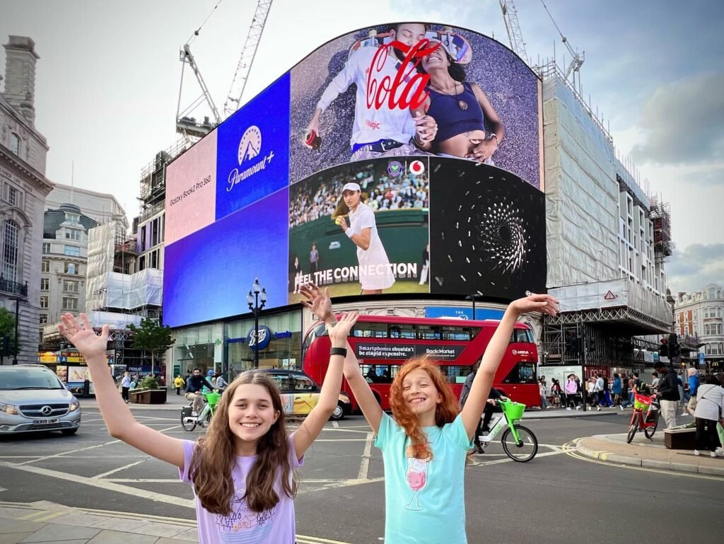 A traveler captures a photo in front of the iconic Coca-Cola clock in London. The image features a bustling street scene with a double-decker bus and large digital screens.