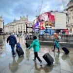 Burghotel “Auf Schönburg” Travelers with rolling luggage crossing a wet European city square with historic architecture and bright street signs.