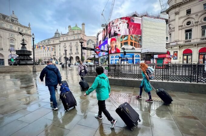 Travelers with rolling luggage crossing a wet European city square with historic architecture and bright street signs.