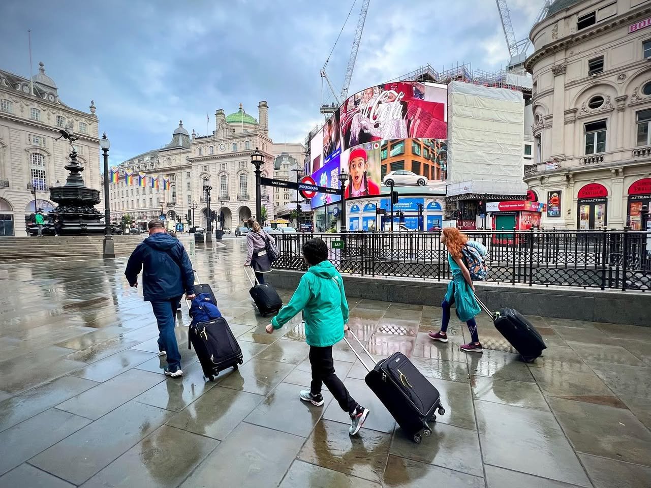 Travelers with rolling luggage crossing a wet European city square with historic architecture and bright street signs.