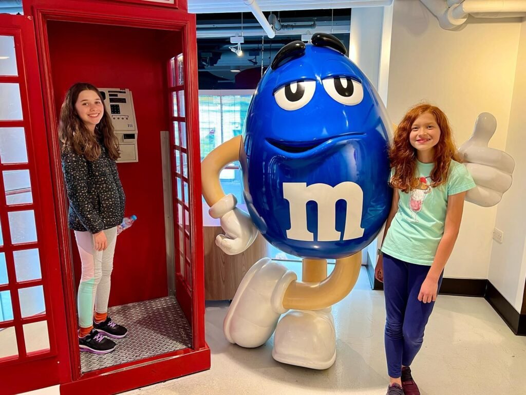 Visitors pose next to a giant red British phone booth and a large blue M&M character in a fun indoor tourist attraction.