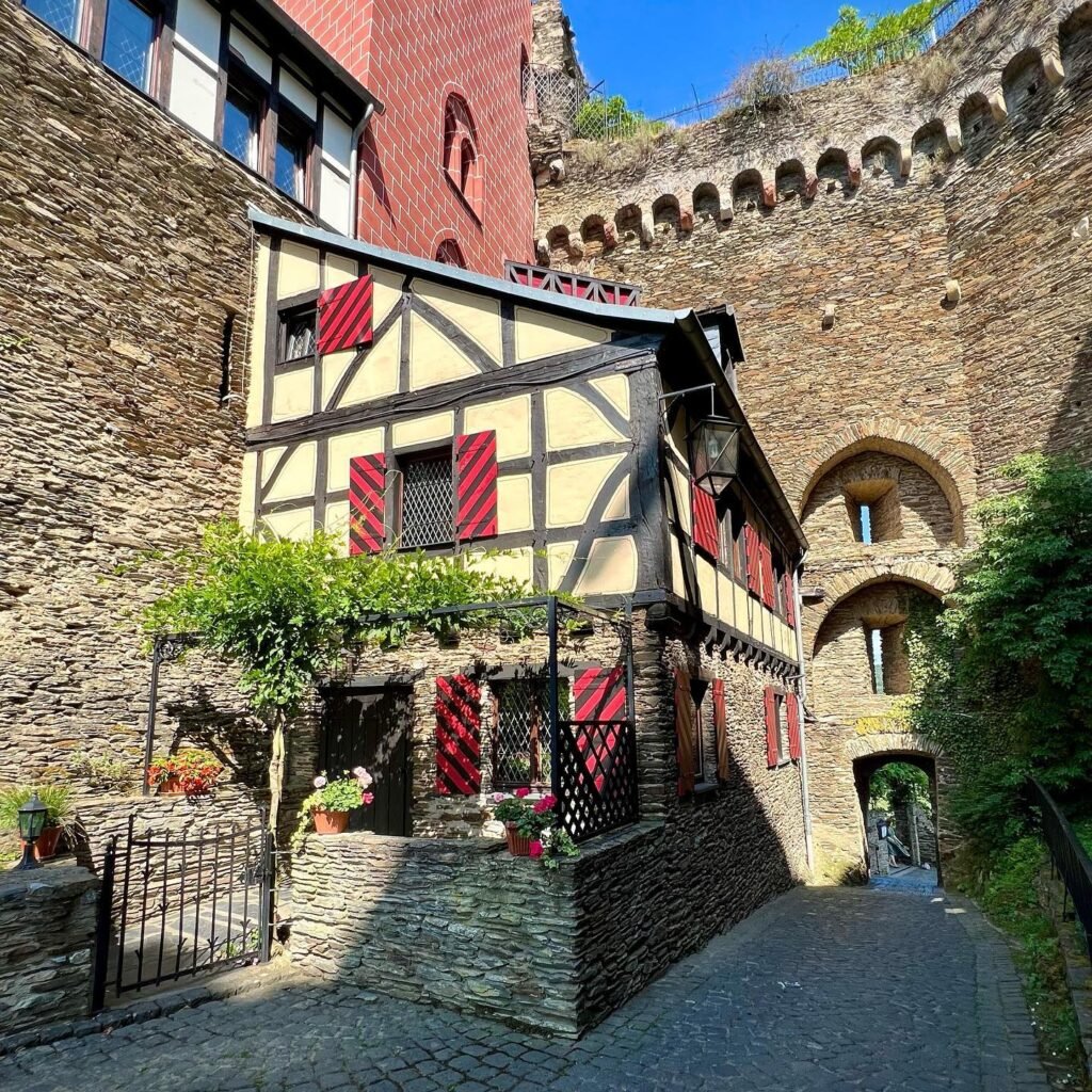 Historic stone castle walls with a charming half-timbered building and red shutters, set on a cobblestone path with arches and greenery under clear blue sky.