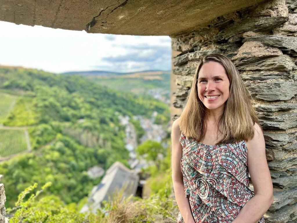 A portrait of a traveler smiling against a rustic stone wall with a lush green valley visible in the background.
