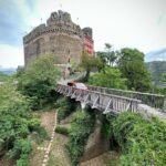 A scenic view of a stone castle with a wooden footbridge leading to its entrance. Lush greenery and a paved pathway surround the historic structure.