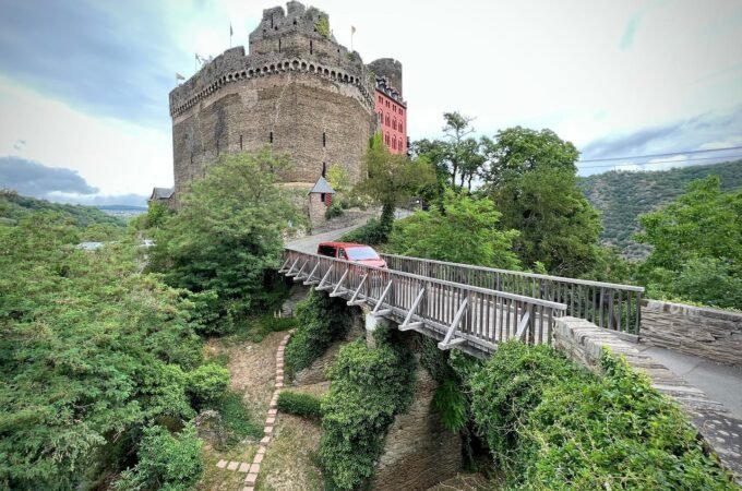 A scenic view of a stone castle with a wooden footbridge leading to its entrance. Lush greenery and a paved pathway surround the historic structure.