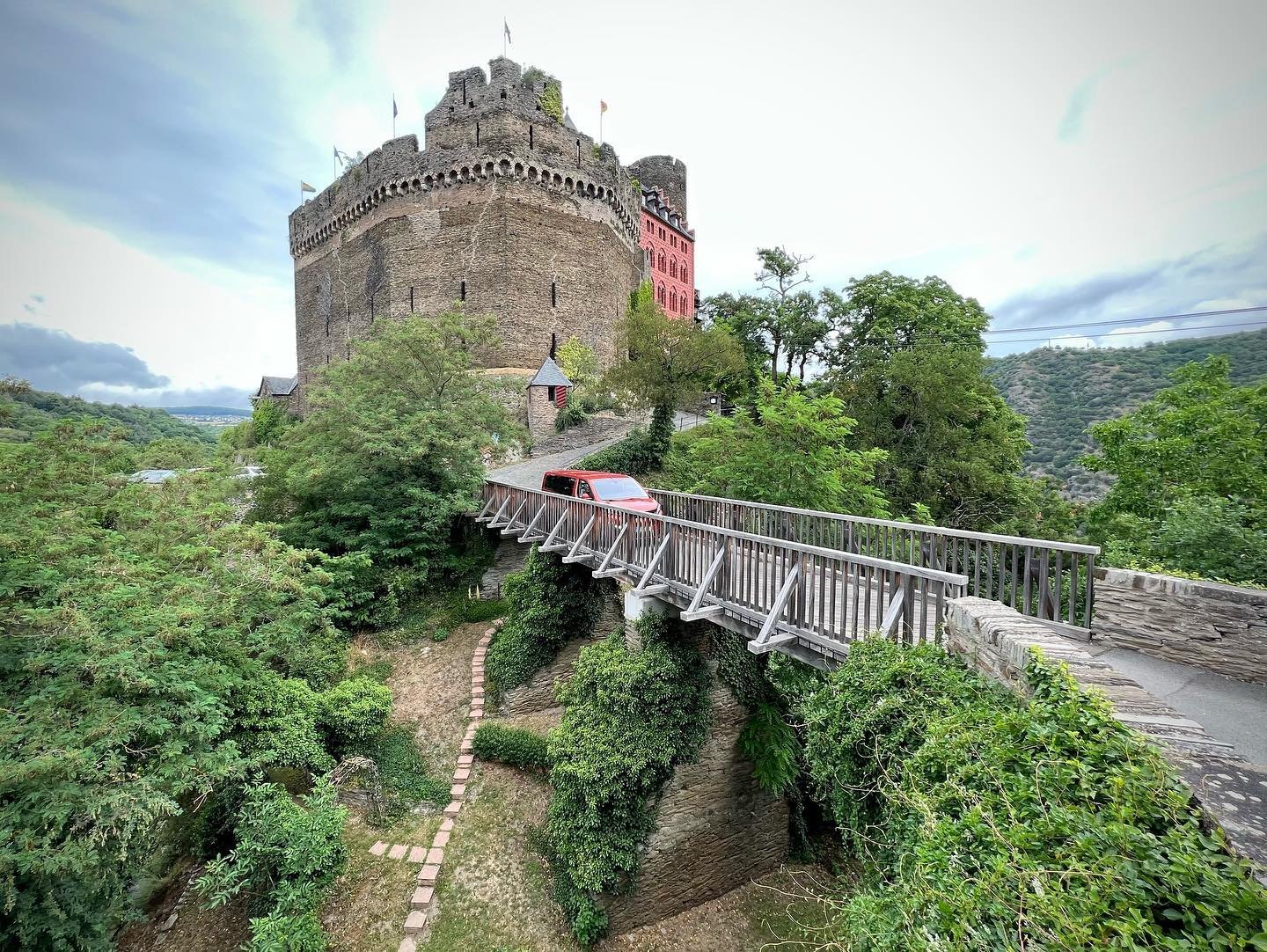 Burghotel “Auf Schönburg” A scenic view of a stone castle with a wooden footbridge leading to its entrance. Lush greenery and a paved pathway surround the historic structure.
