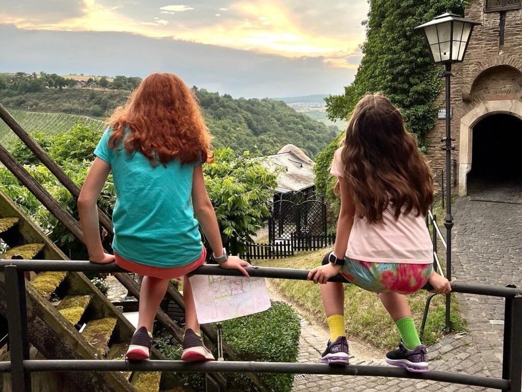 Two children sit on a railing overlooking a lush, hilly landscape. The scene includes a historic stone building and a vintage street lamp.