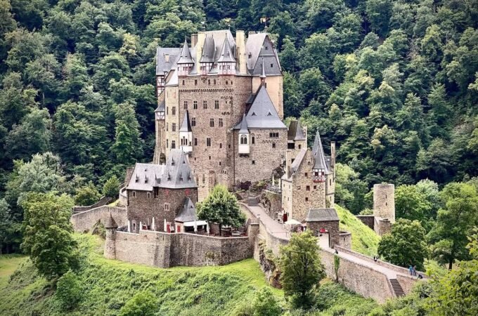 A detailed view of a historic stone castle perched on a lush, green hillside surrounded by dense forest.