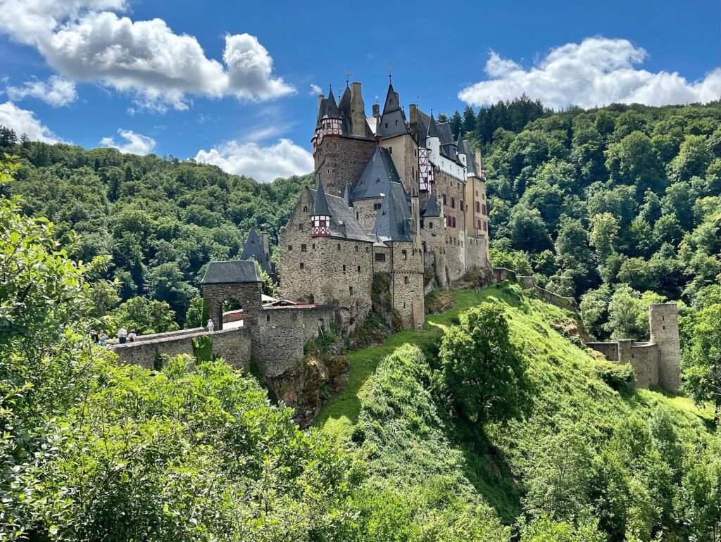 A majestic stone castle nestled in dense green forest under a crisp blue sky. The towers rise dramatically above the hillsides.
