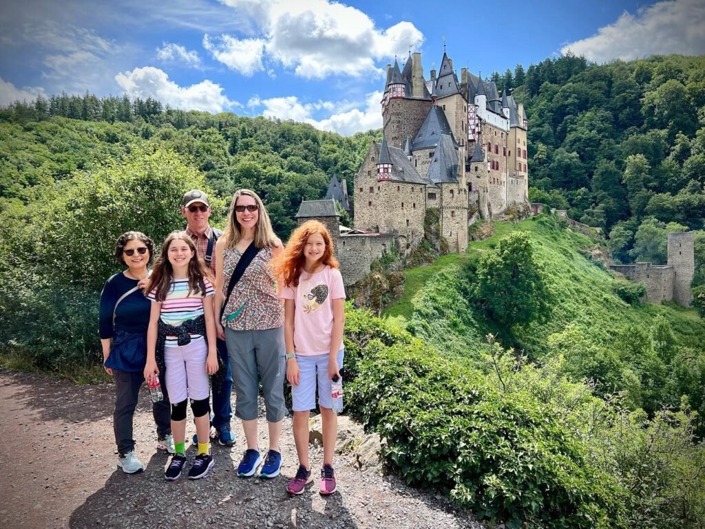 A group poses with the historic Hohenzollern Castle on a hilltop in Germany. Lush forests surround the medieval fortress on a sunny day.