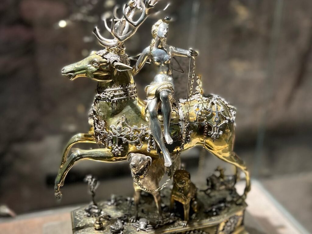 A detailed metal sculpture of a deer with a rider is featured in a series of close-up views. The piece combines rustic textures with polished gold and silver tones.