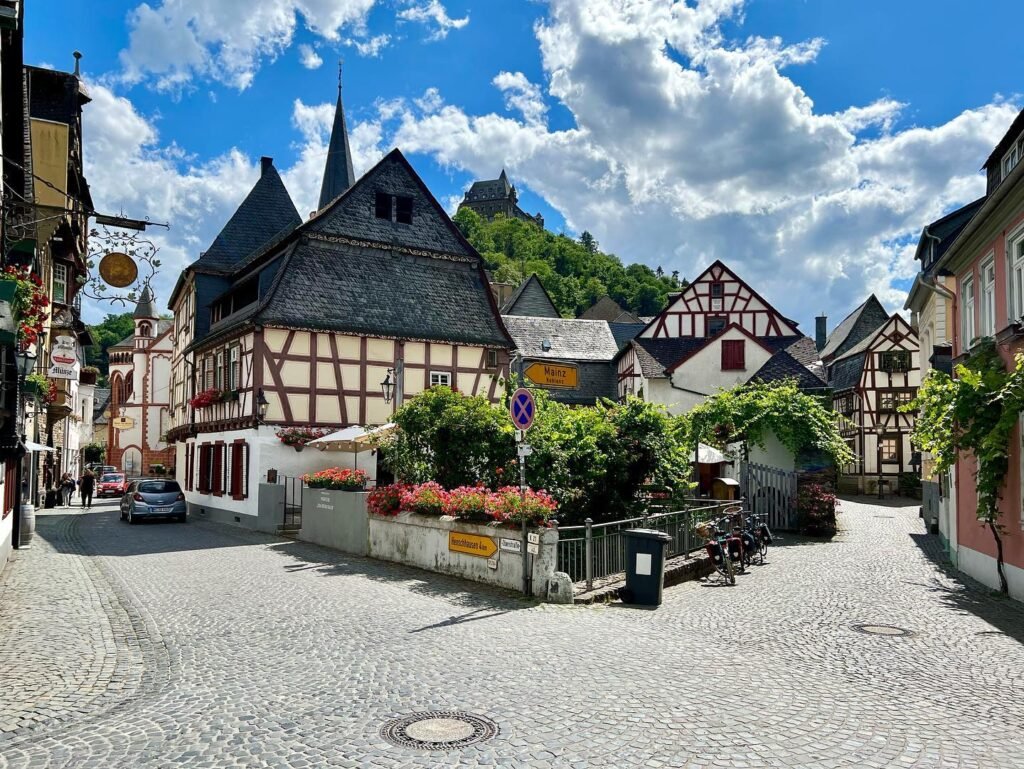 A scenic cobbled street in a European town with timber-framed buildings under a partly cloudy blue sky. A hilltop castle is visible in the background.