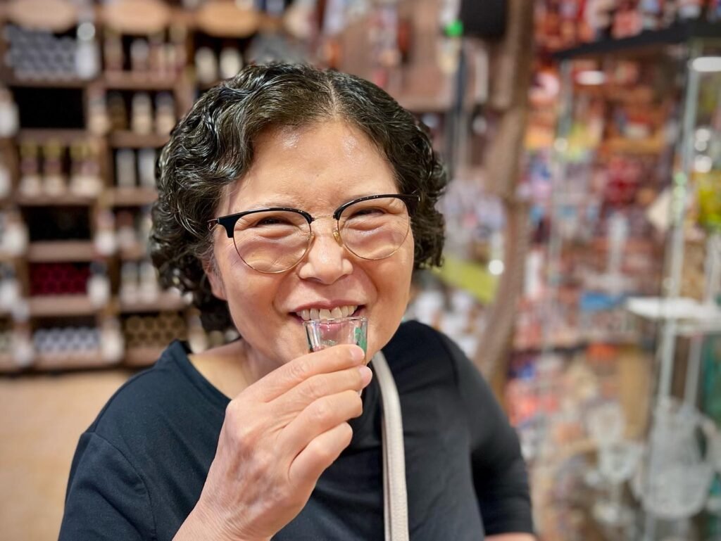 A person smiling while sipping from a small glass inside a shop.