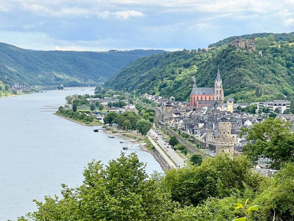 A scenic view of a quaint town nestled along the riverbanks, framed by lush green hills and features a prominent church with twin spires.