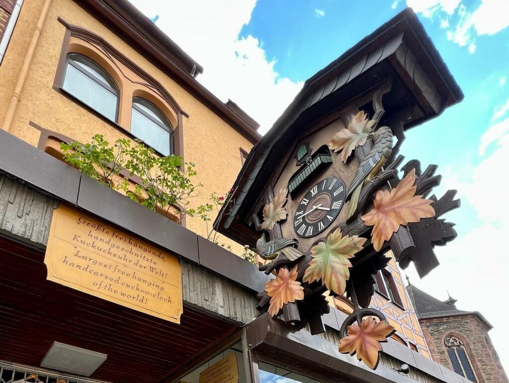 A close-up of a historic clock with decorative leaves and a plaque, set against a beige building with arched windows under a bright blue sky.