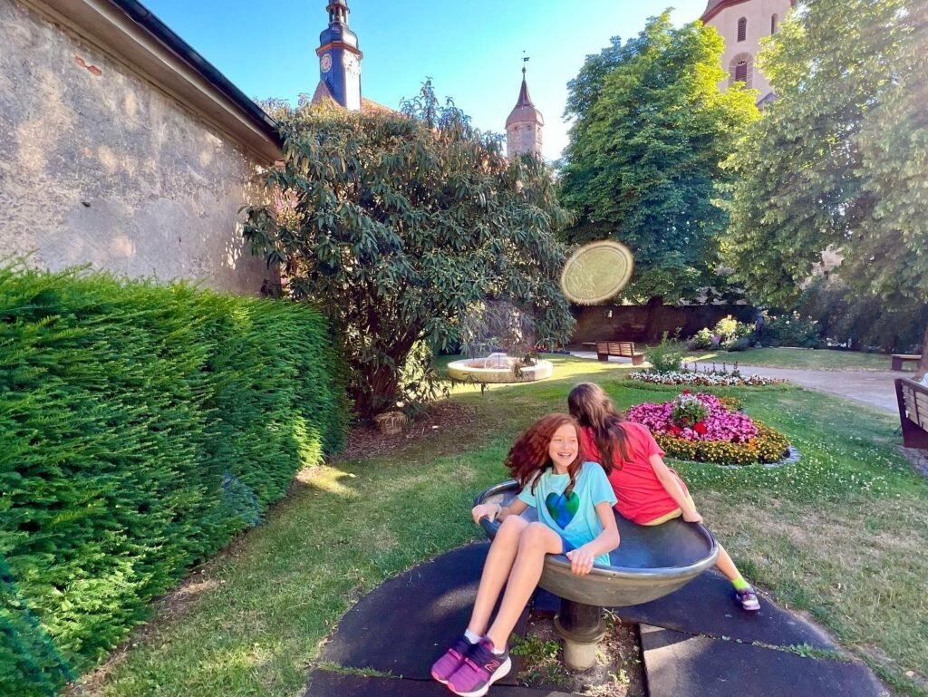 A child and friend sit on a metal bowl structure in a lush garden. Historic architectural elements are visible in the background.