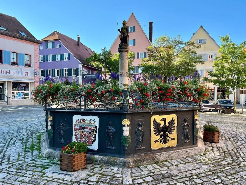 A picturesque European town square featuring a central monument and colorful architectural buildings under a clear blue sky. The cobblestone plaza showcases vibrant flower arrangements and historic charm.