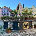 Bacharach A picturesque European town square featuring a central monument and colorful architectural buildings under a clear blue sky. The cobblestone plaza showcases vibrant flower arrangements and historic charm.