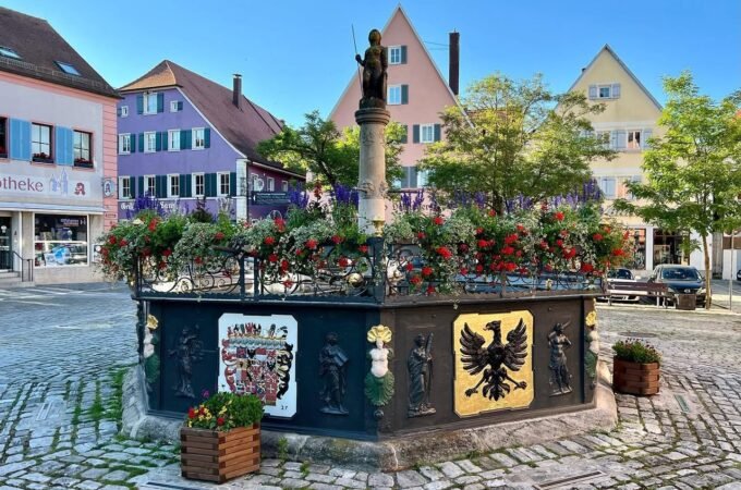 A picturesque European town square featuring a central monument and colorful architectural buildings under a clear blue sky. The cobblestone plaza showcases vibrant flower arrangements and historic charm.