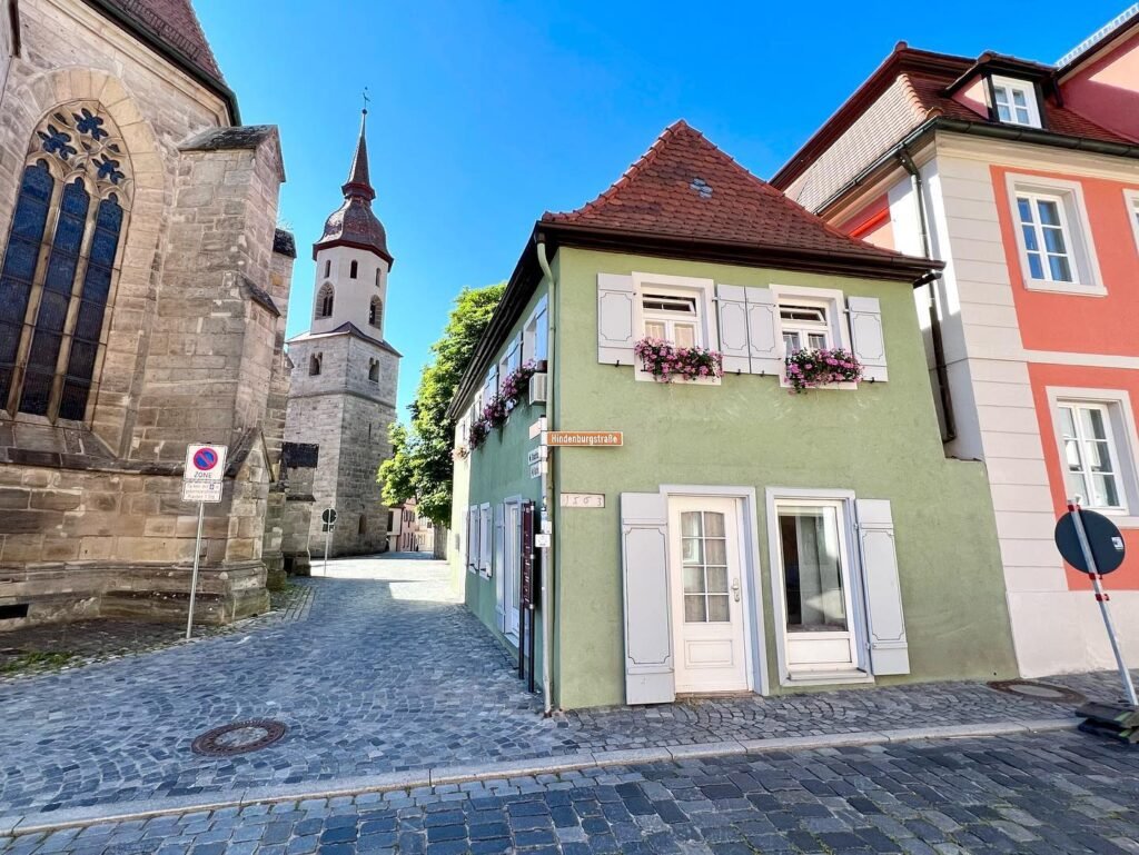 A charming cobblestone street lined with traditional half-timbered buildings, featuring vibrant green and red facades and a historic stone church.