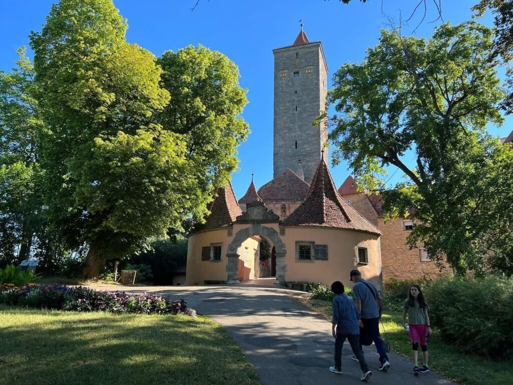 A family walking past a historic stone tower and small church-like building surrounded by greenery.