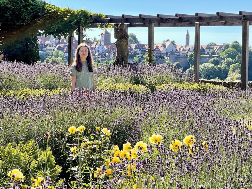A girl is standing in a field of lavender with french countryside in the background.