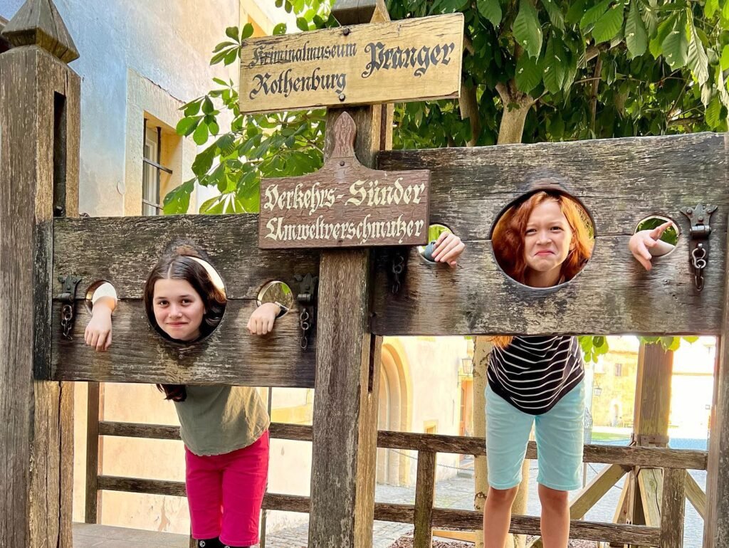 Two individuals peek through holes in a wooden sign at a tourist attraction, with German text visible on the sign.