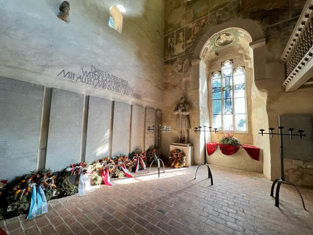 A spacious stone interior with tall windows, flower tributes, and metal stands. Sunlight fills the room with historical architectural details.