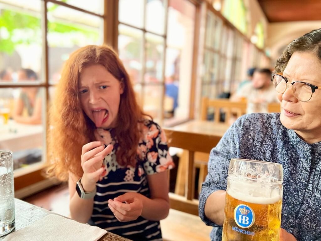 A person makes a playful face while eating at a bright restaurant table next to another person holding a glass of beer.