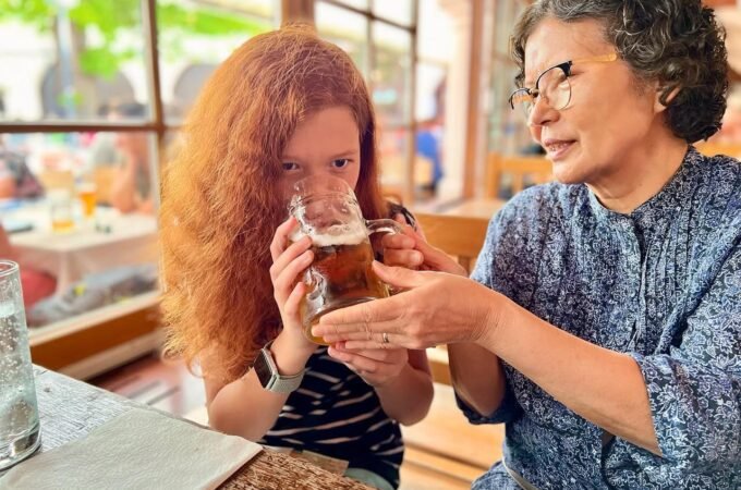 A person with reddish-orange hair sips a drink at a bar while another person stands nearby.