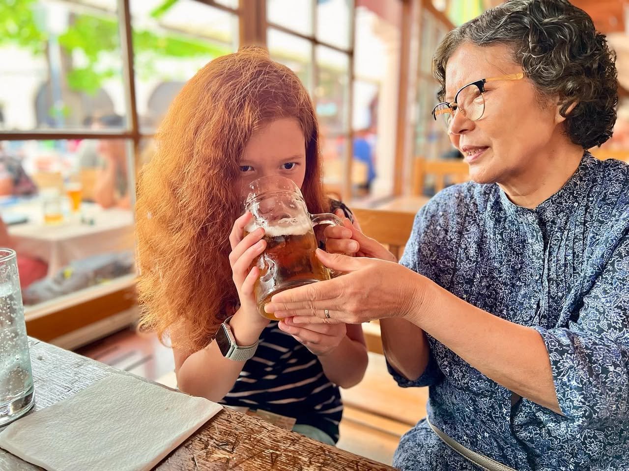 A person with reddish-orange hair sips a drink at a bar while another person stands nearby.
