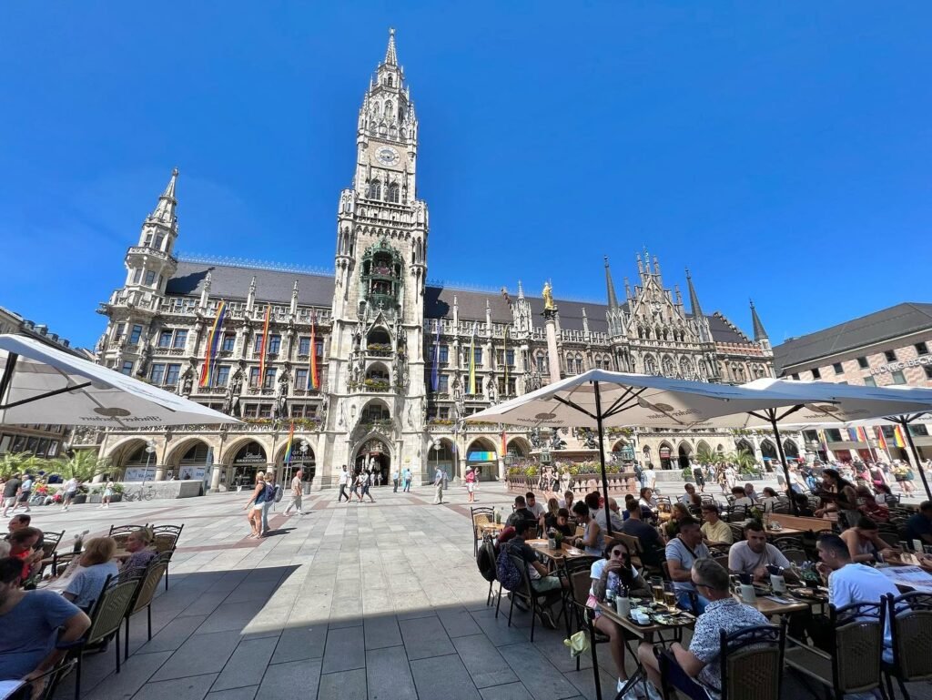 A bustling outdoor café with white umbrellas sits in the courtyard of the Gothic cathedral in Brugge, Belgium. The scene captures a bright, sunny day in the historic square.