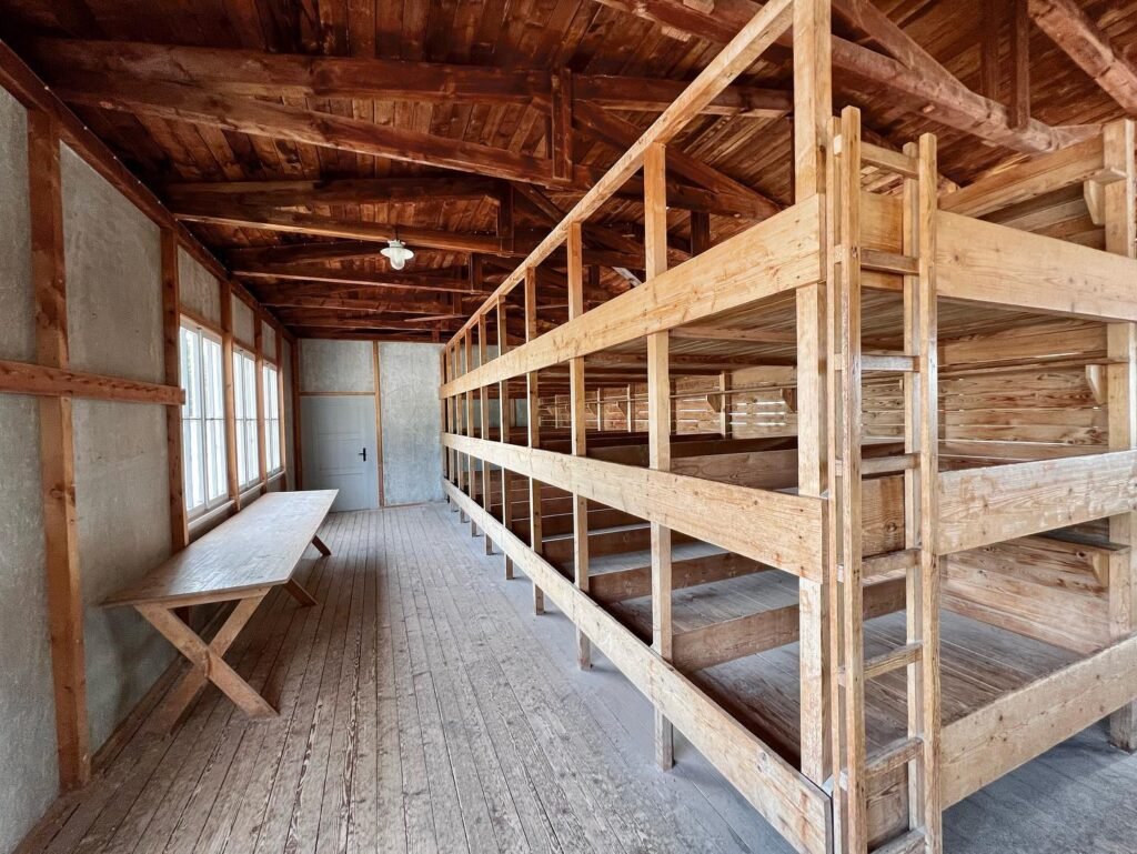 Rows of empty wooden bunks and an old barn ceiling in a rustic dormitory setting, featuring a simple bench under the windows.