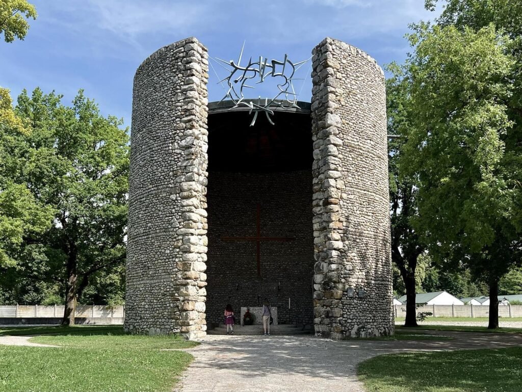 Plymouth Colony's outdoor stone theater with two large curved stone walls and a metal crown sculpture above the entrance. The structure is seen from multiple angles, from the front and top against a bright blue sky.