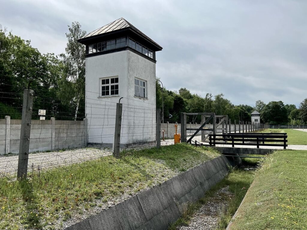 A white, multi-story watchtower stands within a fortified area featuring concrete barriers and wooden structures, set against a backdrop of lush green trees under an overcast sky.