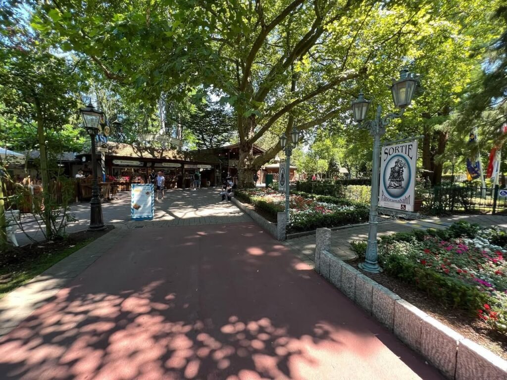 A shaded pathway in a lush green park with vintage street lamps and vibrant flower beds lining the walkway.