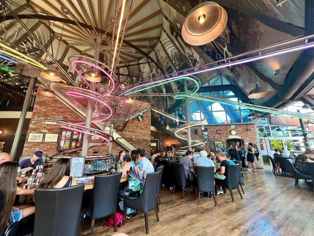 A modern station interior featuring unique neon light installations and a high, industrial ceiling. Rows of seating and patrons are visible in a bustling terminal setting.