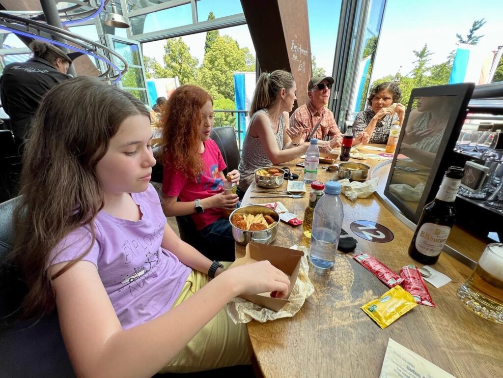 A group enjoys a meal at a sunny, window-lined restaurant. A close-up of a table shows drinks and snacks.