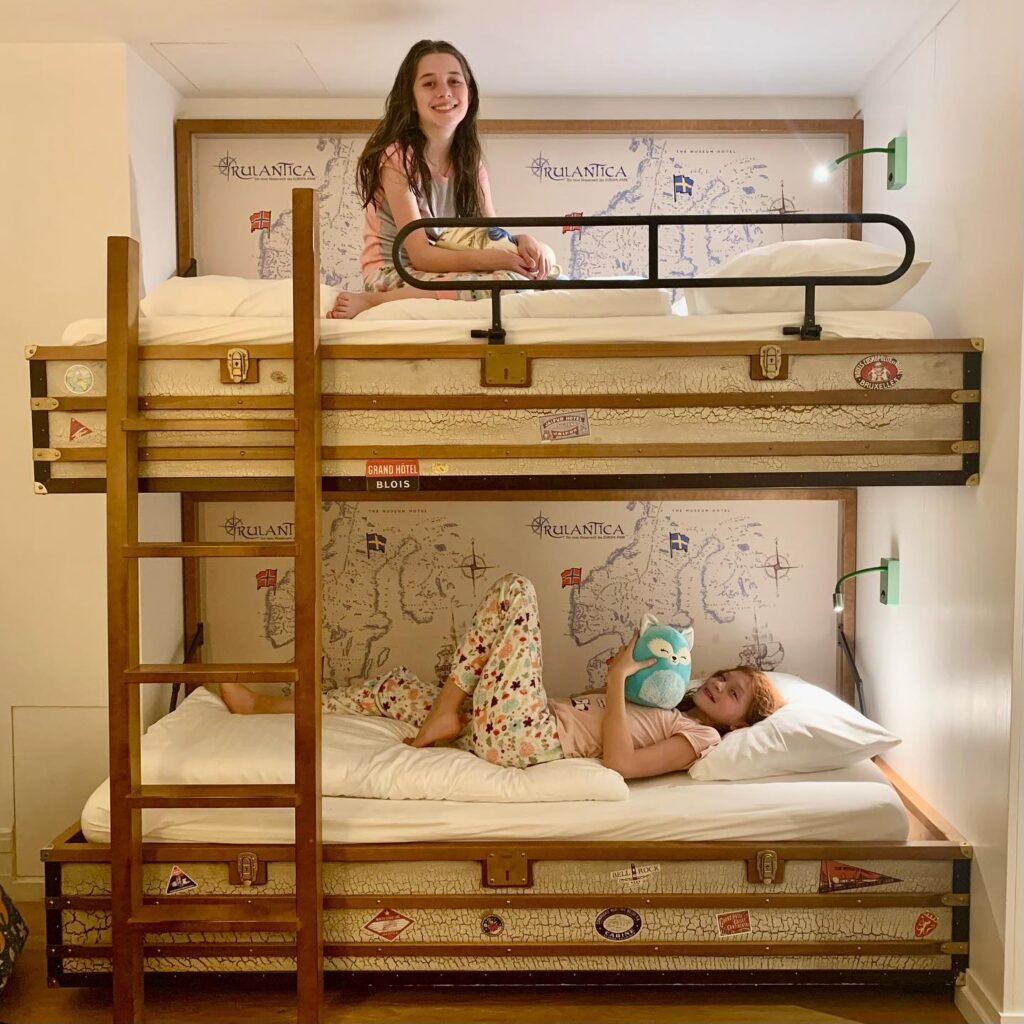 A unique bunk bed constructed from vintage suitcases with travel-themed wallpaper. Smiling visitors relaxing on the beds with a map background.