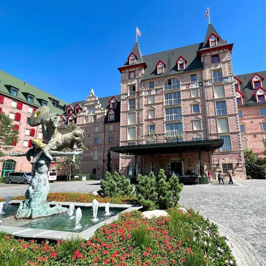 The historic facade of a grand hotel with a decorative fountain and red shutters against a clear blue sky.