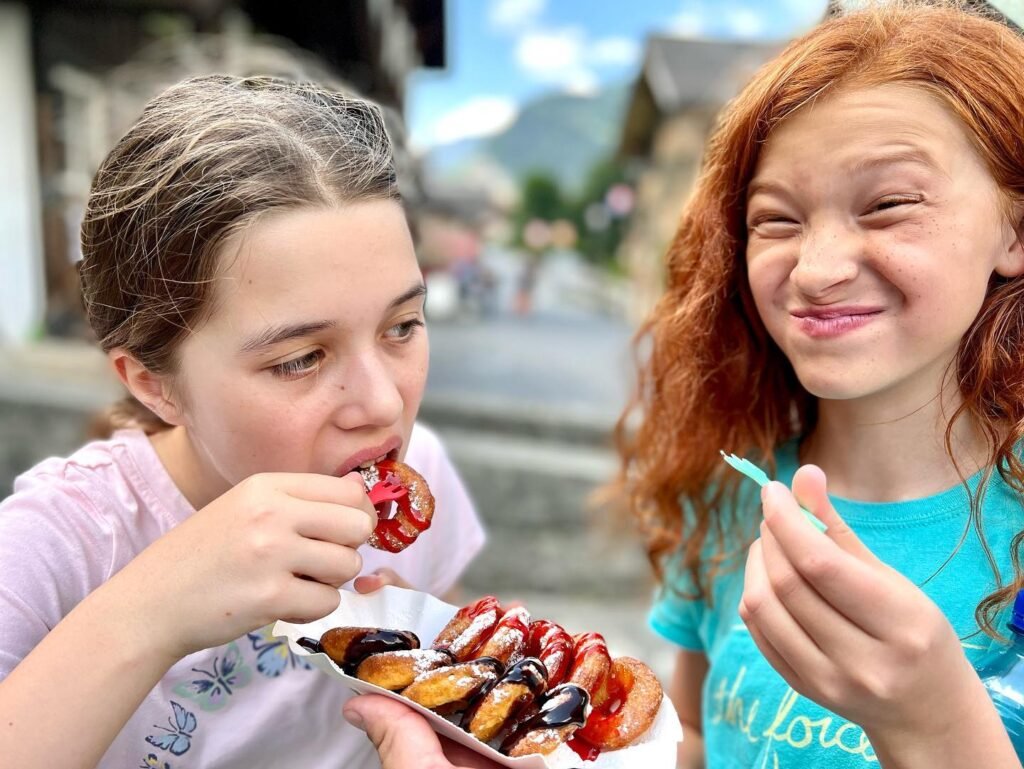 Two children enjoy colorful tommelisa candy from a paper tray in a pedestrian street with historic buildings and a mountain vista.