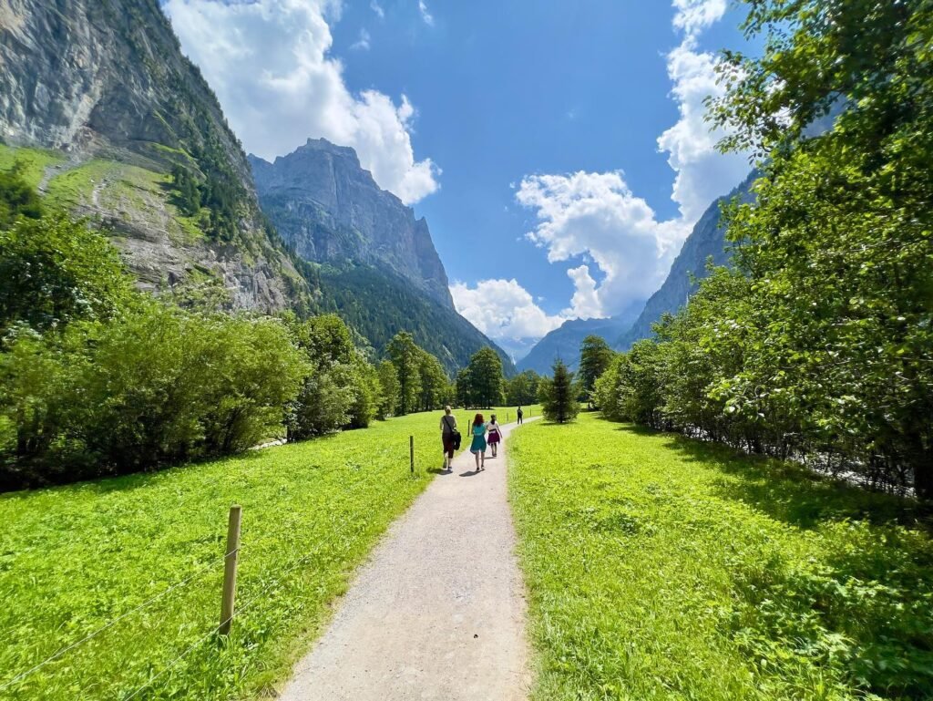 A walking path through the Village Green in Lauterbrunnen surrounded by alpine meadows and steep cliffs. Hikers enjoy the scenery under a bright blue sky with white clouds and lush greenery.
