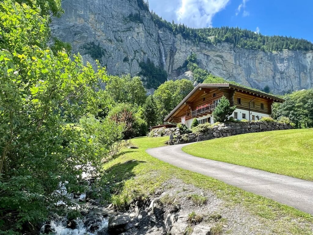 A picturesque scene of a winding road through a lush green valley with a traditional wooden house in the background, framed by majestic mountains and sparkling streams.